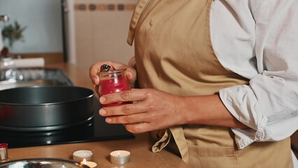 Close-up woman in chef's apron, lighting a candle in cozy home kitchen interior