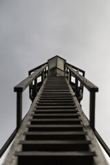 A dramatic upward view of a wooden staircase leading to a lighthouse or beacon, with a cloudy sky in the background, emphasizing perspective and architectural structure.