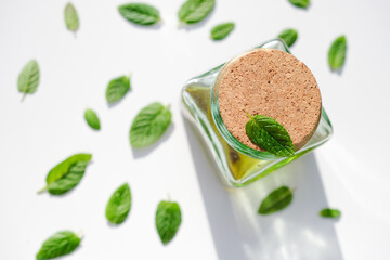 Glass square bottle with yellow-green liquid, with a cork stopper. Mint leaves are scattered across a clean, white surface. Natural wellness, and eco-friendly living concepts. Shallow depth of field.