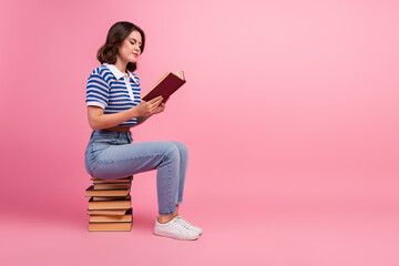 Casual young woman with stylish bob haircut sits on stack of books, reading in tranquil mood on vibrant pink background