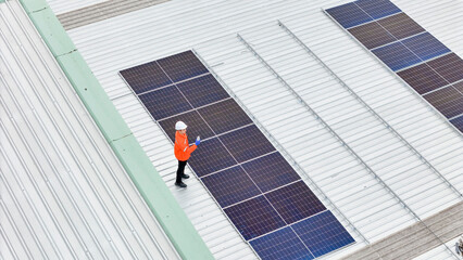 technician in safety gear inspects solar panels on a metal rooftop, holding a tool. The scene...