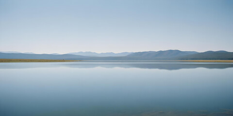 Fototapeta premium A serene lake with calm, reflective waters and distant mountains on the horizon under a clear blue sky. a range of low, rolling hills that appear in soft shades of blue and gray, creating a sense of d