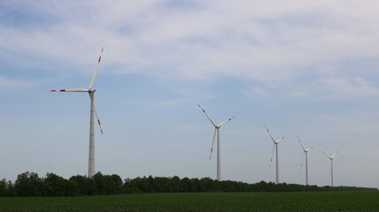 row of wind turbines in green field with underbrush against cloudy sky, industrial technological landscape in natural area, windmills in industrial use