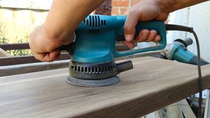 hands holding blue sanding machine and pressing it on the surface of textured wood during polishing process, sanding wood smooth with sanding tool