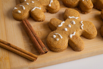 On a bamboo wooden board lie warm, freshly baked gingerbread men, beautifully painted with white icing, with two fragrant cinnamon sticks placed beside them.