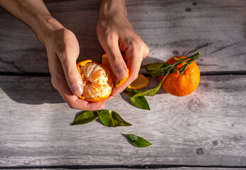 The beautiful hands of a young man are peeling a tangerine on a wooden table, next to the table there is a whole tangerine with a twig and leaves. Close-up.