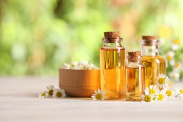 Bottles of essential oil and chamomile flowers on white wooden table against blurred background, closeup. Space for text