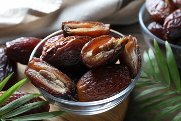 Many tasty dried dates in bowl and leaves on table, closeup