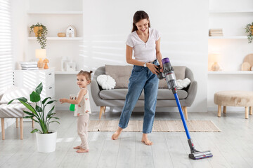 Smiling young woman cleaning floor with cordless vacuum cleaner while her daughter spraying...