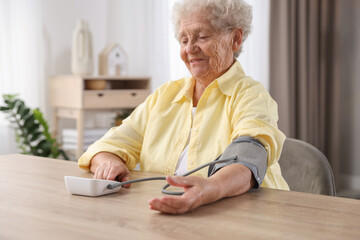 Senior woman measuring blood pressure at wooden table indoors