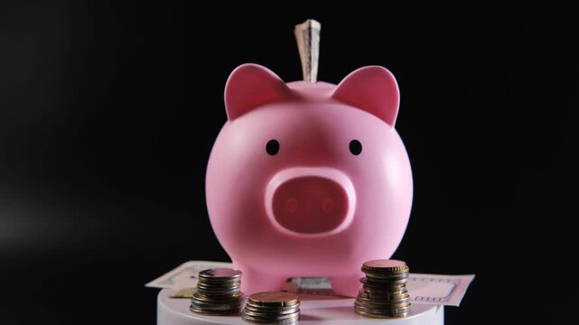 A pink piggy bank spins on a pedestal, surrounded by scattered coins and banknotes, creating a striking contrast with the dark background, evoking savings and financial dreams