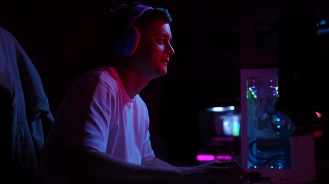 Frustrated young man wearing wireless headphones losing match during playing online video games on powerful computer in dark room illuminated by neon lights. Concept of modern online entertainment.