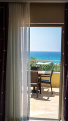 View of Mediterranean Sea through balcony door or window and white curtain. Balcony terrace with table and chairs overlooking the sea. Vertical image