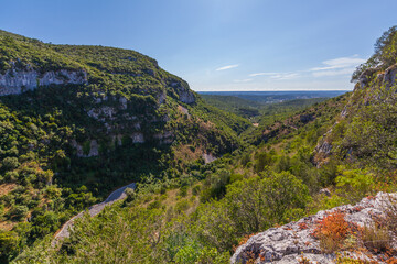 A breathtaking view of a deep valley with steep cliffs, lush vegetation, and a winding road snaking through the landscape. The clear blue sky and distant horizon complete the picturesque scene