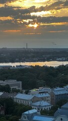 Golden sunset over Riga with warm hues reflecting on the Daugava River. Historic rooftops and lush greenery frame the view, blending urban charm with natural beauty