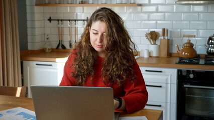 A woman works from home, seated at a table in a modern kitchen. Holding a document while a laptop is open in front of her.