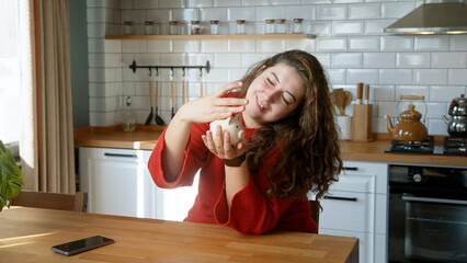 A woman with long curly hair, sits at a table in a modern kitchen while holding a white piggy bank...