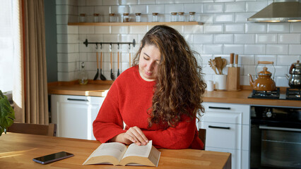A woman with long curly hair, reads a book in a modern kitchen. Focused and relaxed, enjoying her quiet time during the day