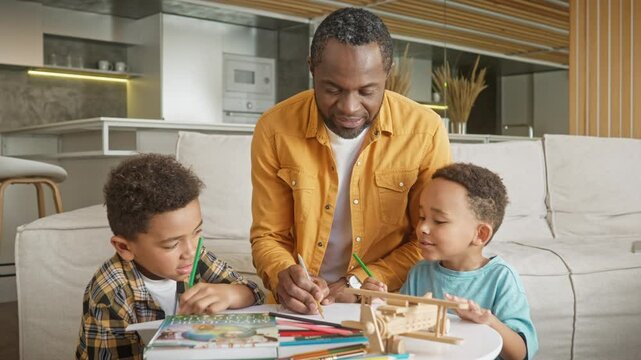 African American father spending some time together with his two kids. Playing with them at table in living room. Teaching kids how to draw by completing illustration together. Education concept.