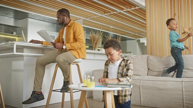 African American man using laptop in living room. Entering data from sheet of paper into system while talking with his colleagues via earphones. Active children playing with toys near busy father.