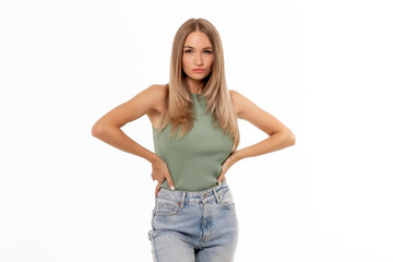 Fototapeta premium A confident young woman poses with hands on hips, wearing a sleeveless green top and distressed jeans against a plain background