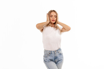 A young woman in a white top stands with her hands over her ears, appearing pensive against a plain...