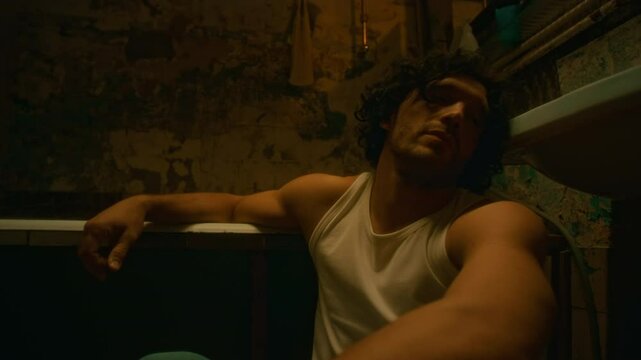 Chest up lowkey shot of exhausted man experiencing pain and escaping reality sitting alone on floor in ruined bathroom in evening, clinging onto sadness