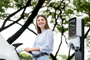 Young woman recharging battery for electric car during road trip travel EV car in natural forest or...