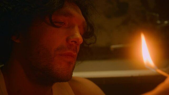 Side lowkey closeup of bearded apathetic Caucasian man striking match and watching it burn while sitting alone in shabby bathroom in evening, questioning meaning of life