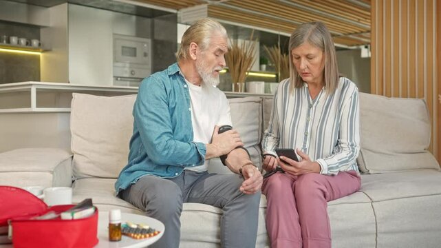 Pretty Caucasian female measuring pressure of her lovely husband in living room. Woman using medical tonometer while her lovedperson feeling bad. Taking care about sick man. Concept of health care.