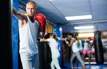 Determined active man practicing boxing punches in gloves on hanging punching bag in training room..