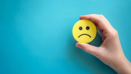 Sad face emoji on a blue background. A hand holds a sad face emoji on a bright blue background. Negative and dissatisfied emotions, bad customer reviews