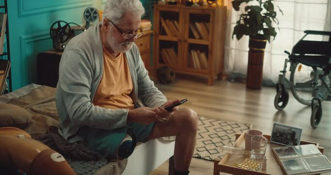 Old 70s man with an amputated leg sitting on bed in his room at home and using a mobile phone, scrolling pages and typing on touch screen