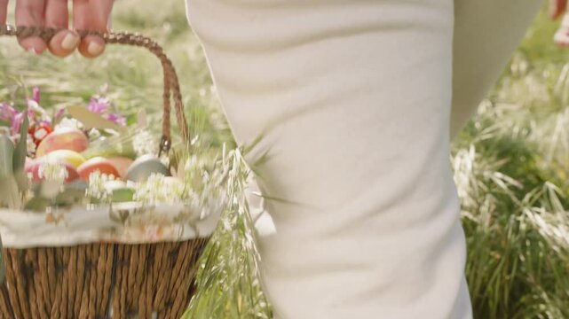 A Woman Walks Away, Carrying a Basket of Colorful Easter Eggs and Flowers Through a Sunlit Field.