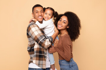 Fototapeta premium Happy family. Cheerful black parents holding their little daughter on hands, embracing and posing together over beige studio background, smiling at camera