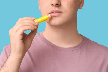 Young man applying lip balm on blue background, closeup