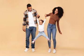 Naklejka premium Excited African American parents playing with their little daughter, girl holding hands with mom and dad and jumping up, family having fun over beige background