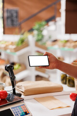 Mobile phone with a white screen held up above a counter in a grocery store. Salesperson horizontally grasping smartphone displaying an isolated chromakey mockup template.