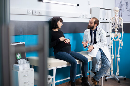 Bearded doctor explains healthcare treatments to young expectant mother during routine maternity checkup. Pregnant woman gently caresses belly bump as obstetrician provides prenatal care advice.