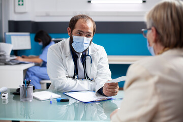 Masked doctor with face mask giving prescription paper to elderly woman in medical office. Female pensioner patient receiving written healthcare treatment plan from young general practitioner.