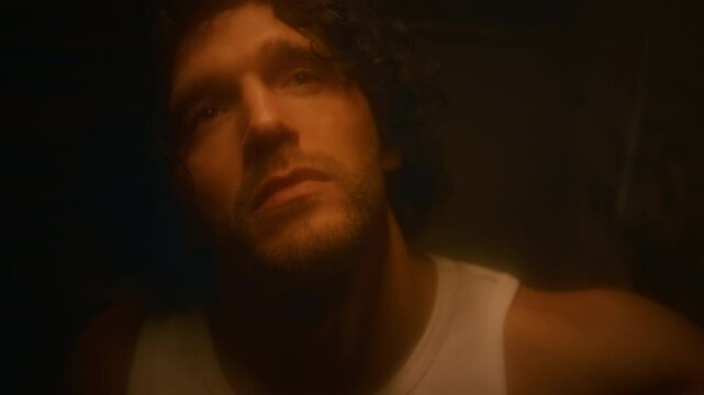 Closeup portrait of unhappy Caucasian man with stubble in undershirt wiping matted glass and looking into camera, feeling depressed and despondent