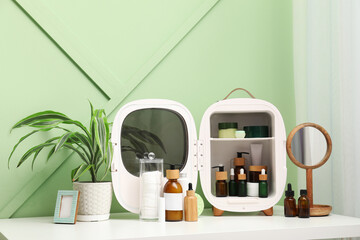 Small refrigerator with cosmetic products, houseplant and mirror on table against green wall
