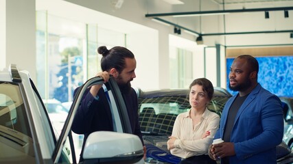 Dealership salesman opening car door, explaining various aspects of vehicle, such as make, model, mileage rates and features. Showroom agent helps customers evaluate potential automobiles, camera A