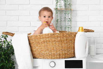 Cute little baby sitting in laundry basket with clothes and bottle of detergent