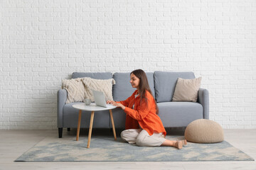 Young pretty woman using laptop on floor near grey sofa at home