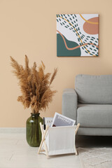 Painting hanging on beige wall, comfortable sofa, vase with pampas grass and magazine holder in room