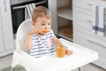 Cute little baby with jar of tasty food eating on chair in kitchen at home