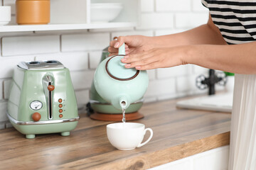 Beautiful young woman pouring hot water from teapot into cup in kitchen, closeup