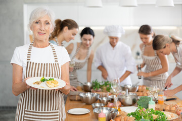 Elderly woman poses with prepared chicken dish at cooking master class