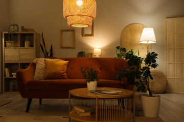 Modern stylish living room with orange sofa, houseplants and glowing lamps in evening